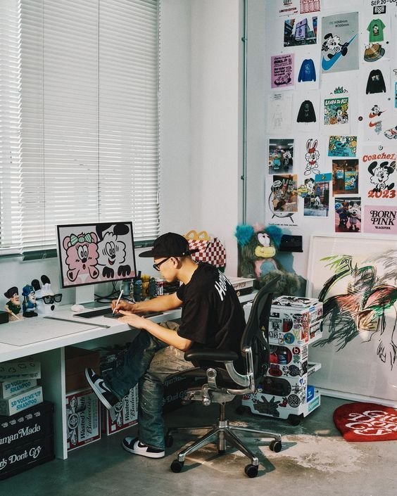 An artist working at a white desk in a creative studio filled with pop-culture posters, collectibles, and stickers