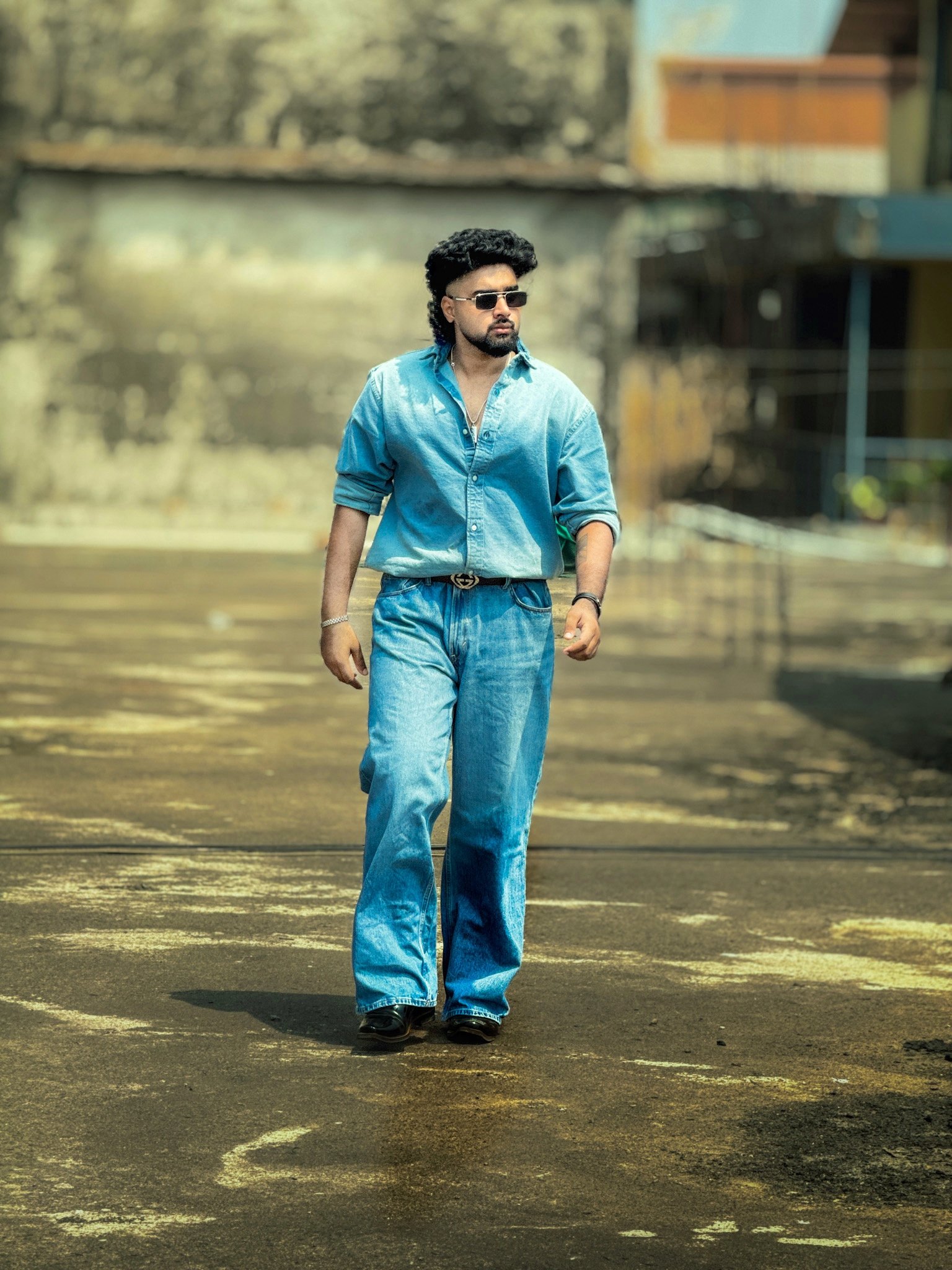 A man with a mullet hairstyle, beard, and sunglasses walks forward in a full denim outfit, featuring a light blue button graphic designer -down shirt tucked into wide-leg jeans. He is outdoors in a sunlit, industrial-looking paved area with a blurred background.